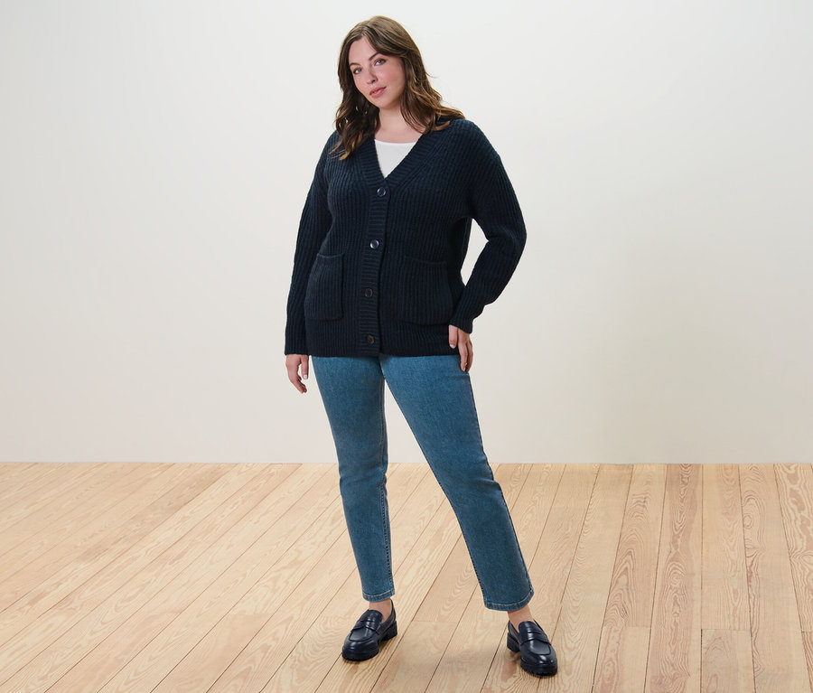 Une femme debout porte un cardigan en grosse maille côtelée bleu foncé, un t-shirt manches 3/4 blanc et un jean droit.