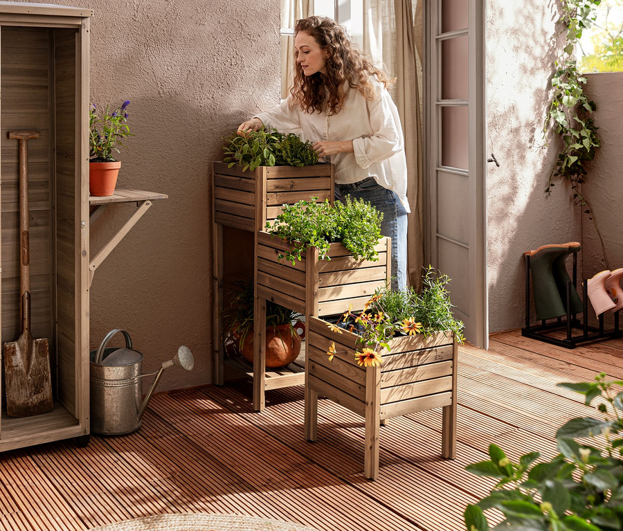 Sur une terrasse, une femme se tient près d'une jardinière en escalier « Tinus » avec étagère, à côté d'une grande armoire de jardin « Tinus » avec étagère latérale rabattable.