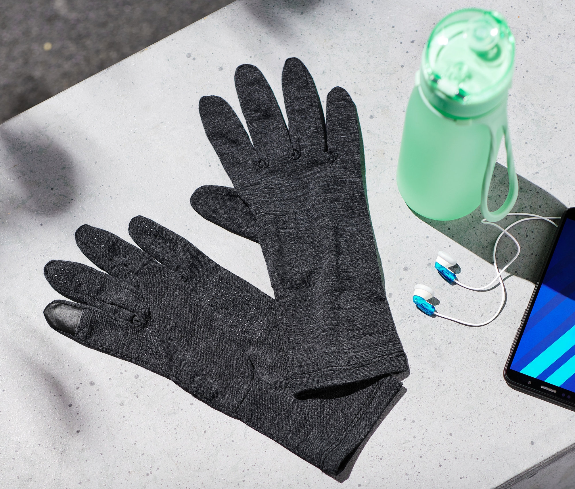 Sur une surface grise reposent des gants en mérinos, une bouteille d'eau, des écouteurs et un téléphone portable.