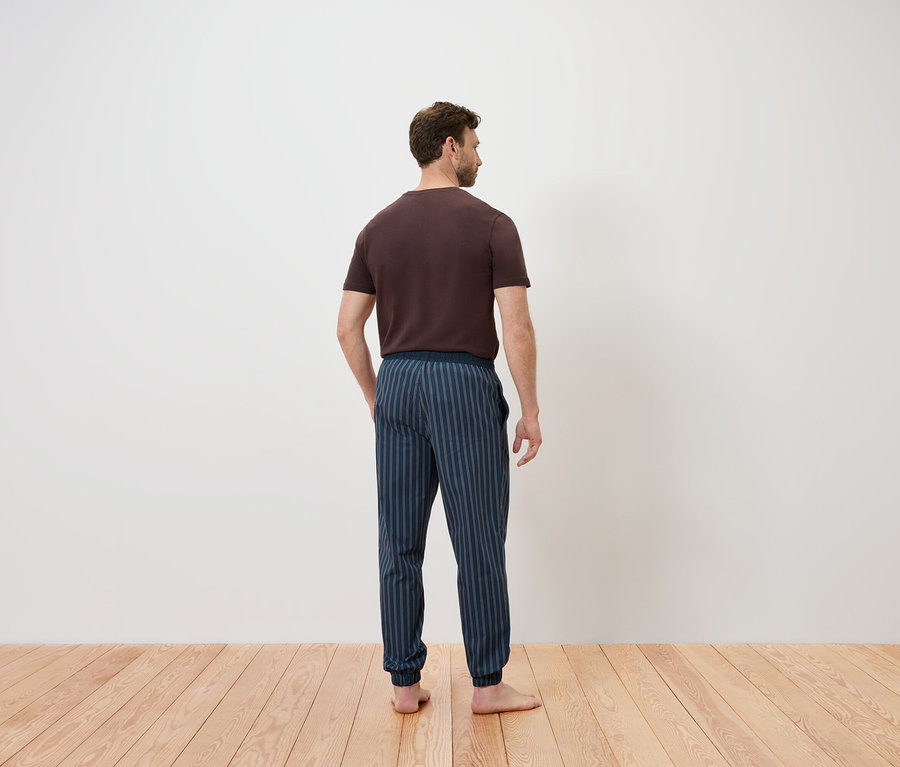 Un homme, dos à la caméra, se tient sur un plancher en bois. Il porte un t-shirt marron et un pantalon rayé bleu.