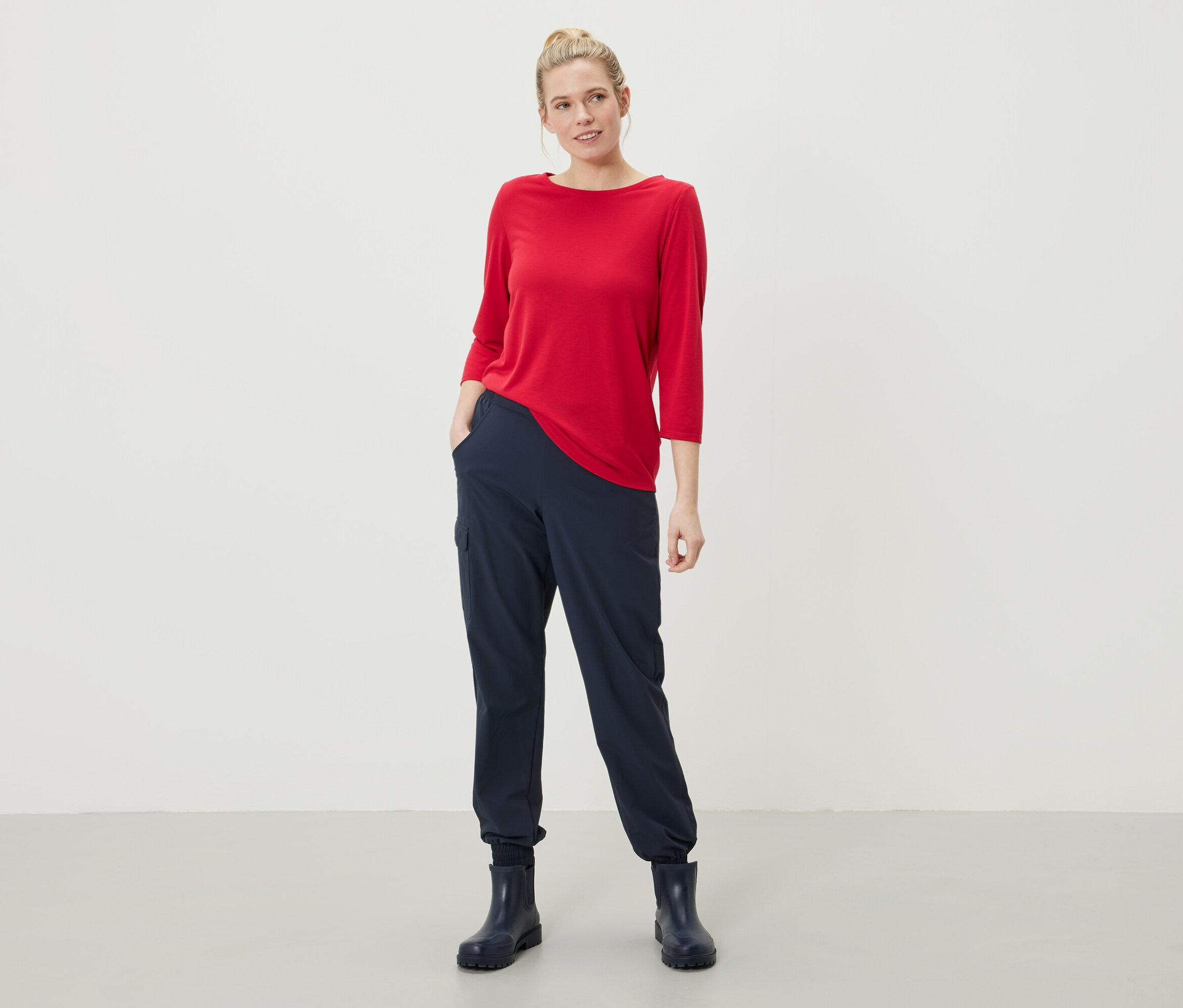 Une femme se tient debout, les mains dans les poches de son pantalon bleu marine, portant un haut rouge à manches trois-quarts et des bottes en caoutchouc bleu marine.
