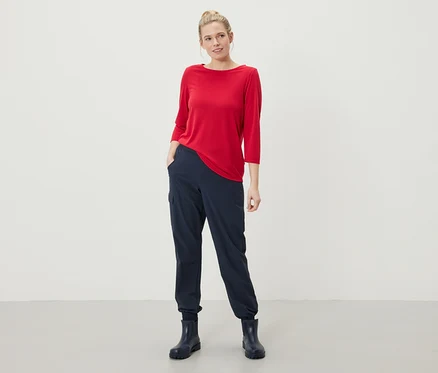 Une femme se tient debout, les mains dans les poches de son pantalon bleu marine, portant un haut rouge à manches trois-quarts et des bottes en caoutchouc bleu marine.
