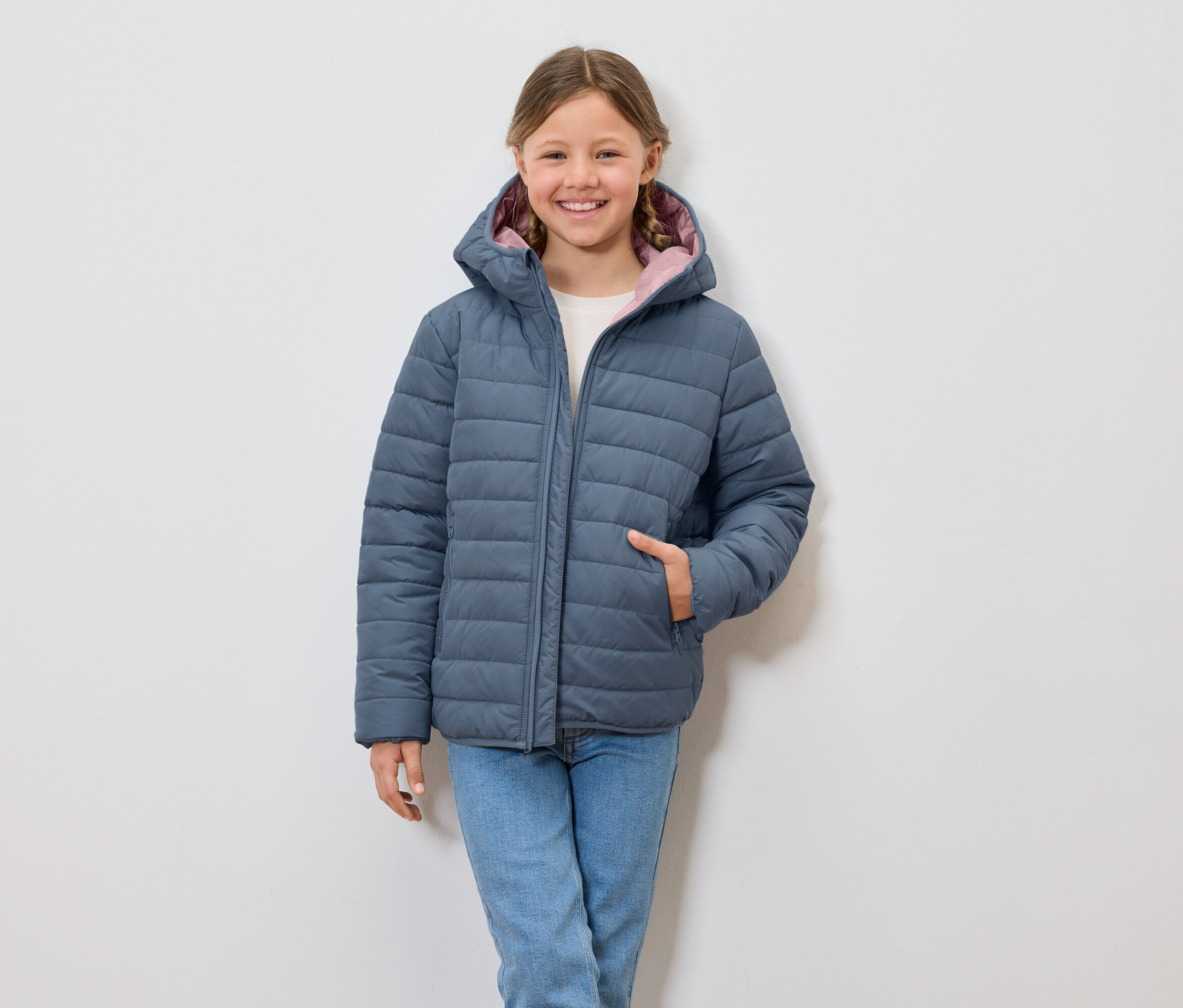 Une fille souriante porte une veste matelassée bleue à capuche et un jean.