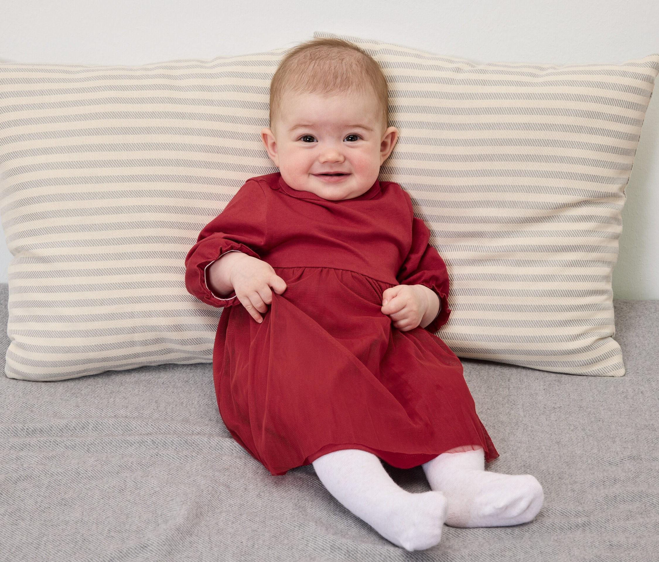 Un bébé souriant en robe rouge est assis sur une couverture grise avec un oreiller rayé.