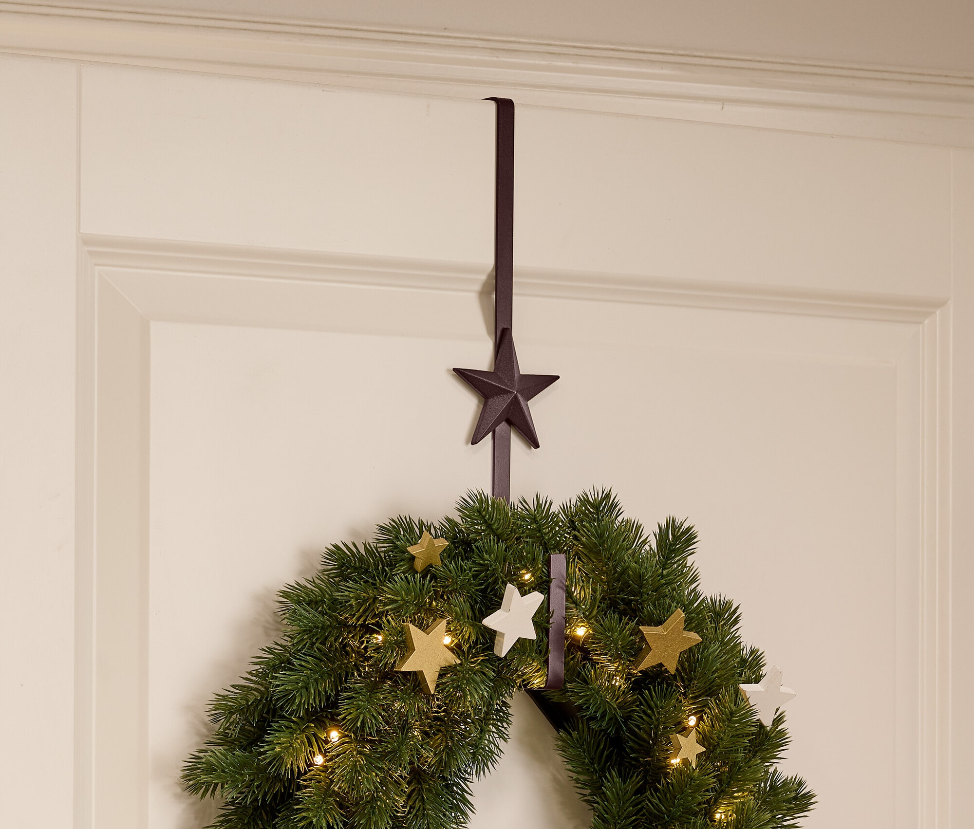 Une couronne de Noël verte avec des étoiles et des lumières suspendue à une porte blanche à l'aide d'un cintre marron en forme d'étoile.