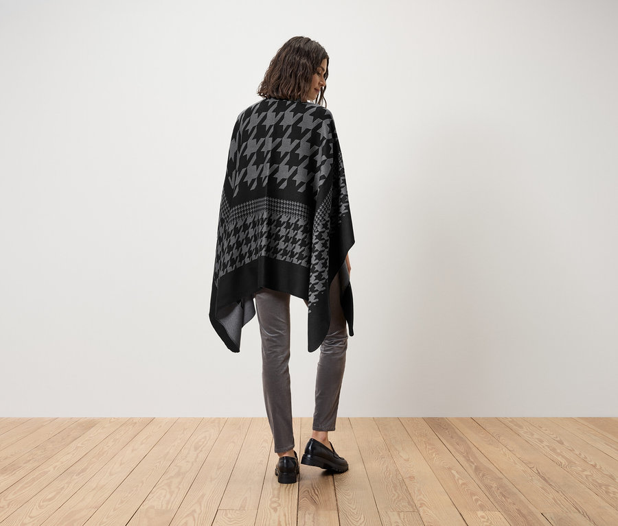 Une femme porte un poncho pied-de-poule gris et noir, un pantalon gris et des chaussures noires.