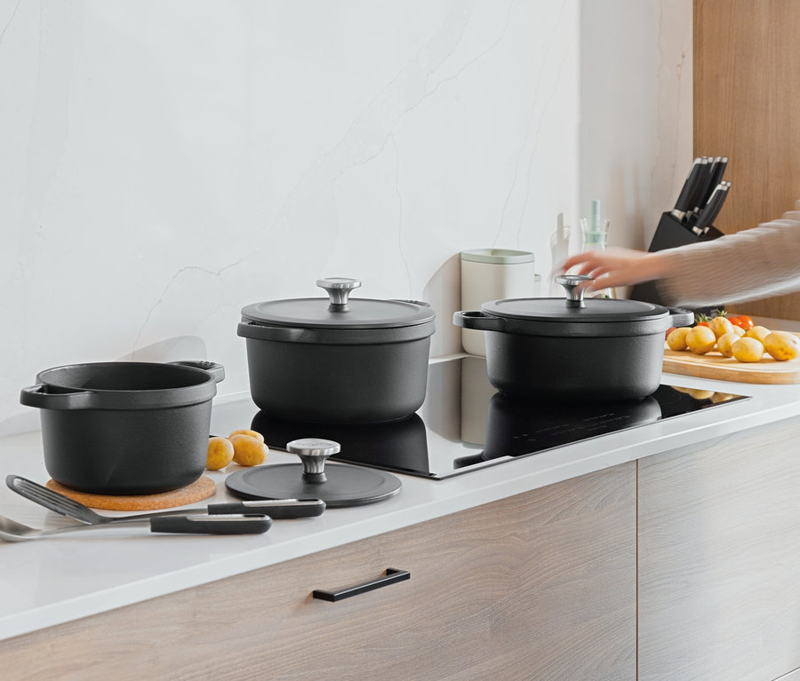 Sur un comptoir de cuisine, trois casseroles noires sont posées à côté de pommes de terre et de tomates. Une main s'approche du couvercle d'une des casseroles.