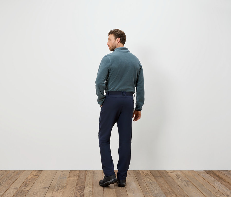 Un homme en pantalon bleu et pull en polaire se tient dos à la caméra sur un plancher en bois.