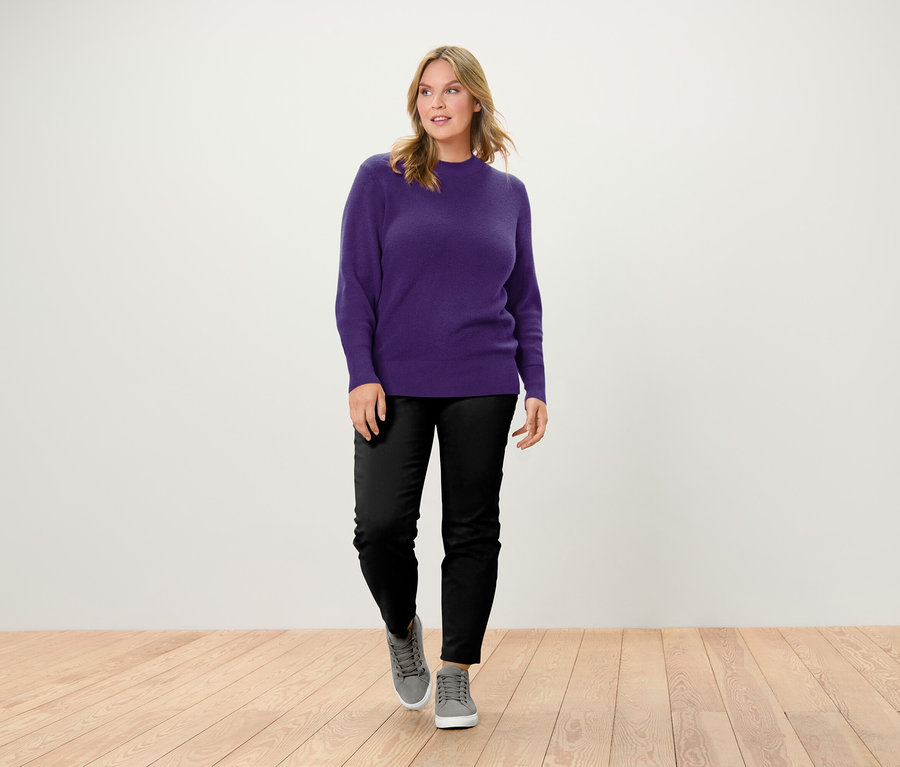 Une femme marche sur un plancher en bois, portant un pull violet, un pantalon noir et des baskets grises.