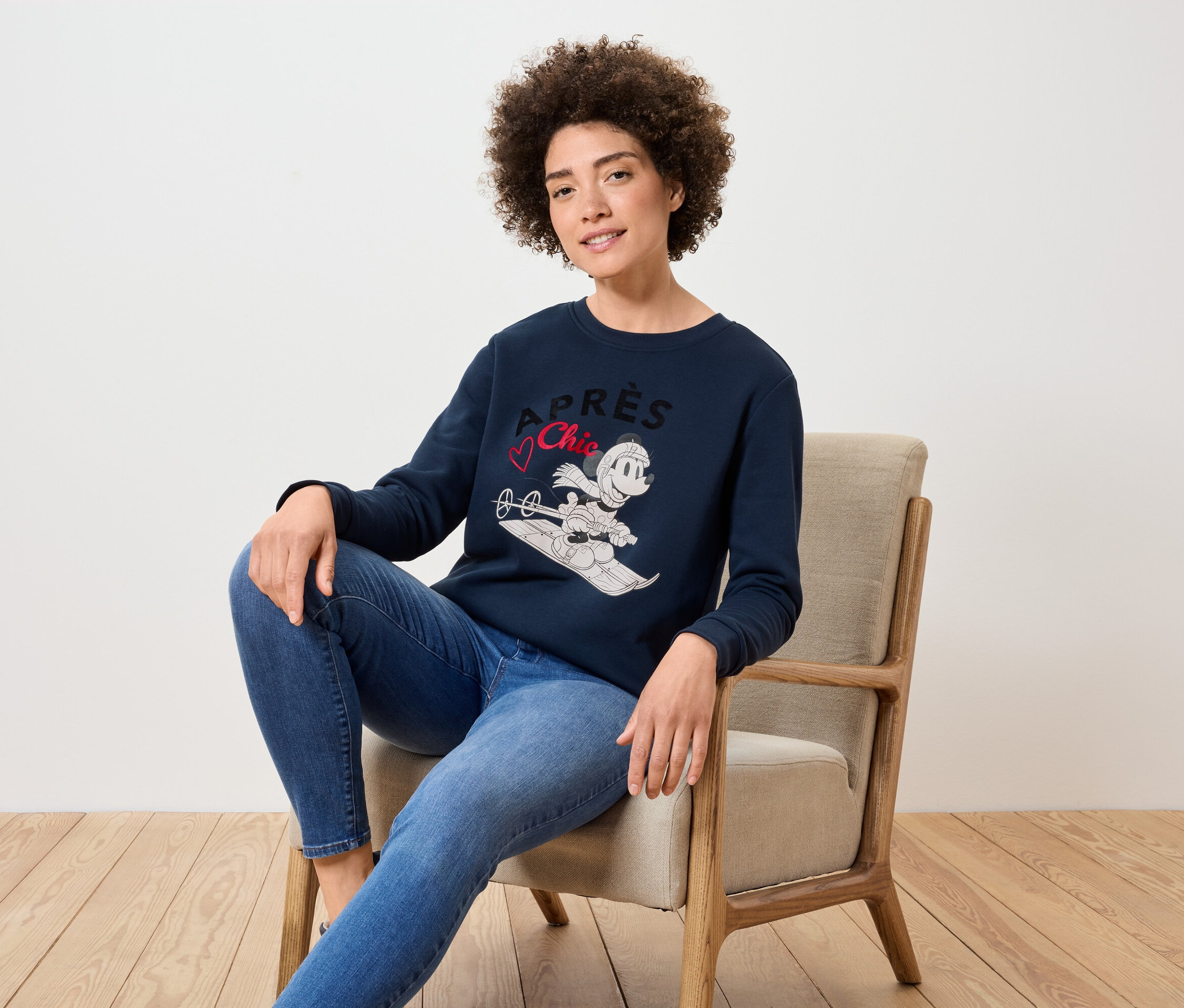Une femme est assise dans un fauteuil et porte un pull bleu marine avec une image de Mickey Mouse faisant du ski et un jean bleu.