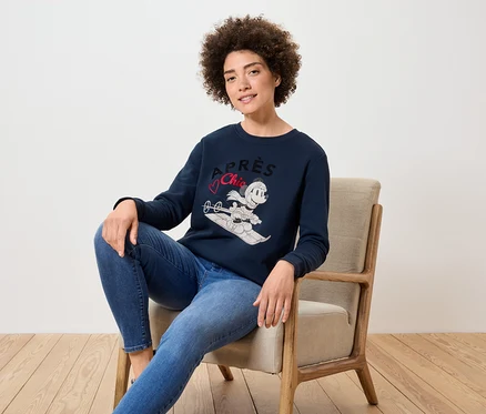 Une femme est assise dans un fauteuil et porte un pull bleu marine avec une image de Mickey Mouse faisant du ski et un jean bleu.