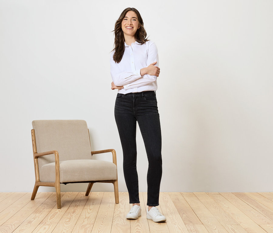 Une femme se tient debout, les bras croisés, à côté d'un fauteuil beige, portant un jean noir et une chemise blanche.