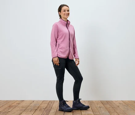 Une femme portant une veste en polaire rose, un pantalon noir et des bottes se tient sur un plancher en bois.