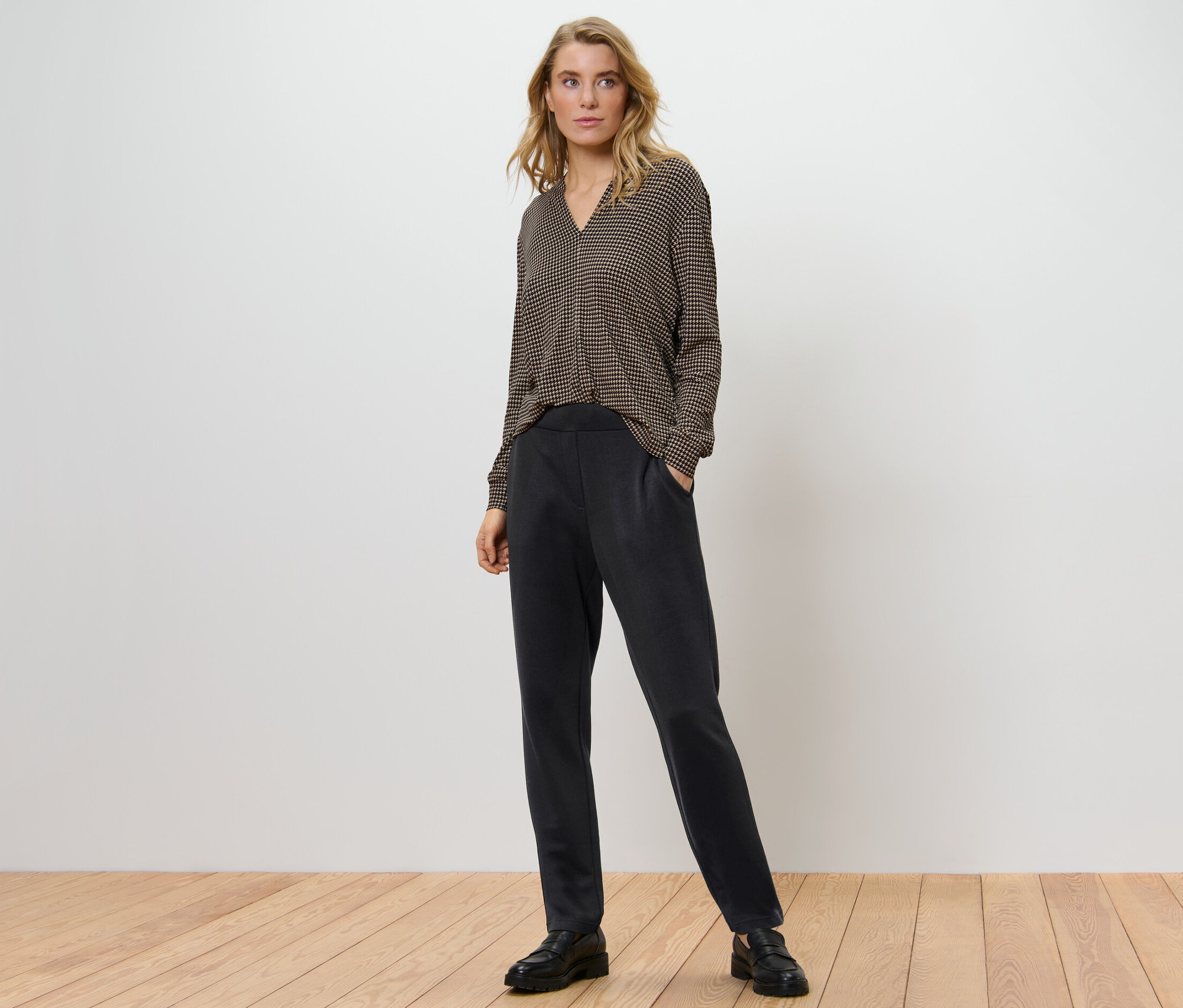 Une femme pose, portant un pantalon de jogging noir et un chemisier à motifs.