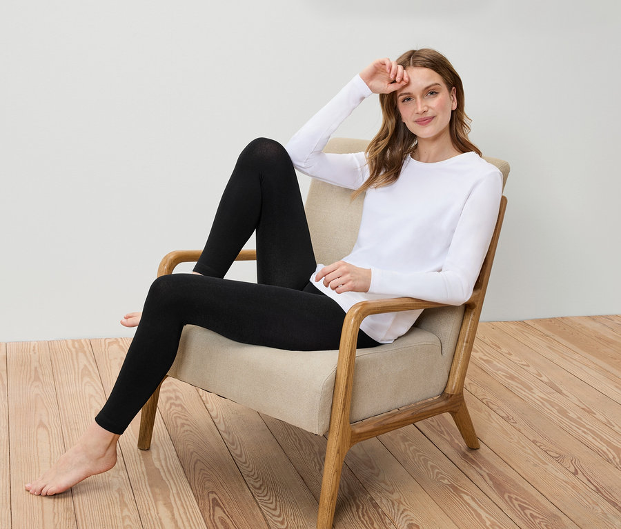 Une femme est assise dans un fauteuil, portant un t-shirt blanc à manches longues et un legging noir.