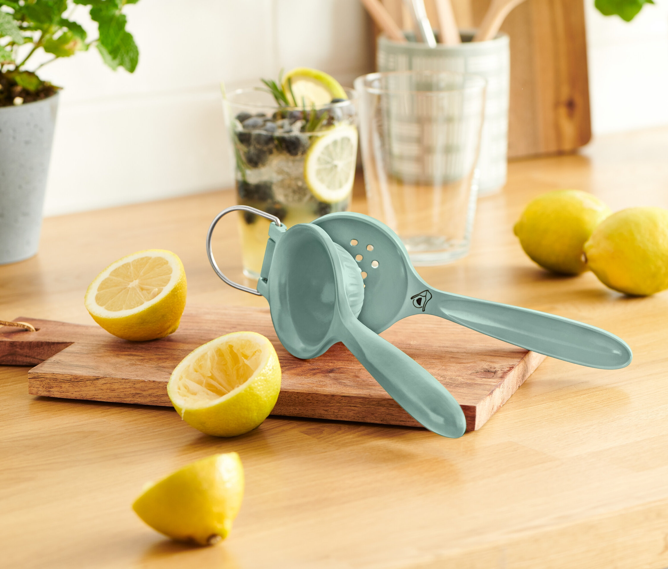 Un presse-citron bleu repose sur une planche à découper en bois à côté de citrons coupés en deux. Un verre avec du citron et des myrtilles est visible en arrière-plan.