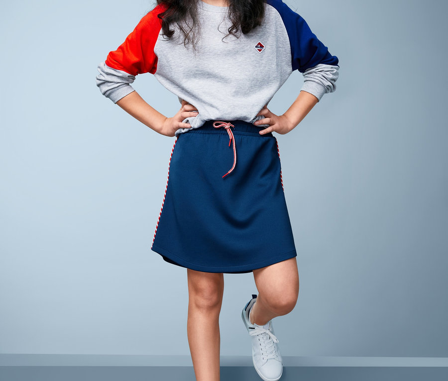 Une fille se tient les mains sur les hanches et porte une jupe sportive en jersey et un pull à manches colorées.