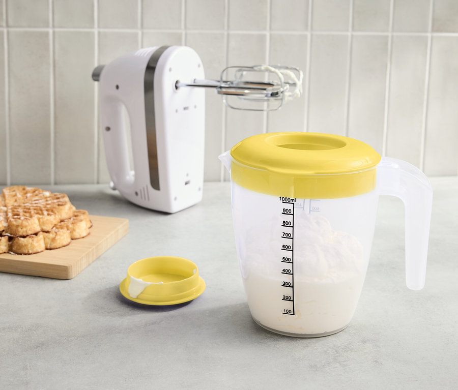 Sur le plan de travail, un mixeur blanc, un verre doseur avec un couvercle jaune rempli de liquide et une pile de gaufres sur une planche.
