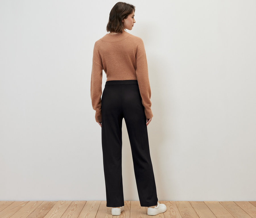 Une femme se tient dos à la caméra et porte un pantalon en jersey avec passepoil noir, un pull marron et des baskets blanches.