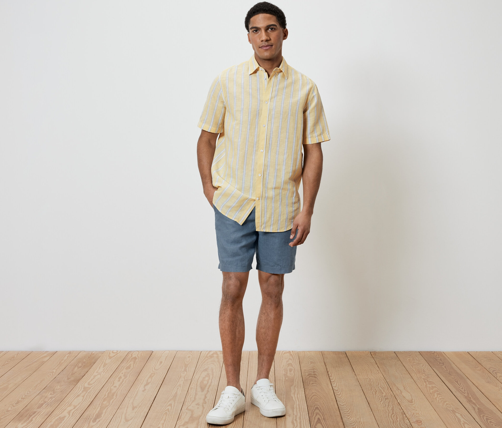 Un homme debout portant une chemise à manches courtes en lin jaune et un short bleu.