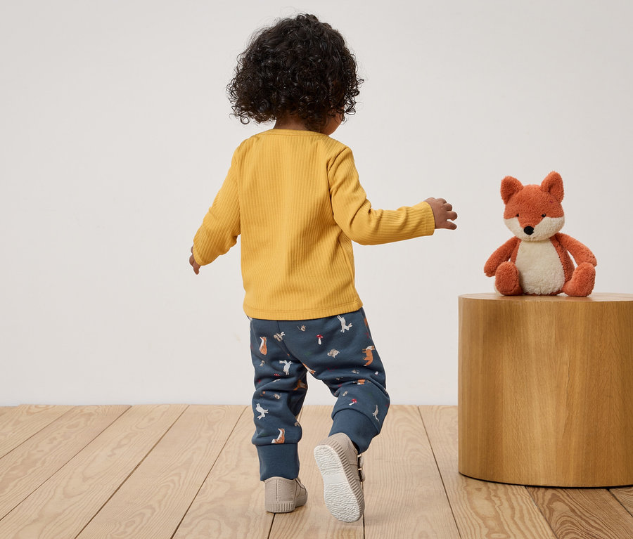 Un enfant aux cheveux bouclés, portant un t-shirt jaune et un pantalon à motifs, marche vers un jouet renard.