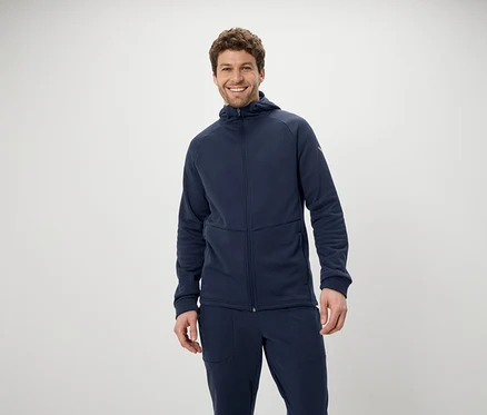 Un homme souriant pose en survêtement bleu à capuche et pantalon sur fond blanc.