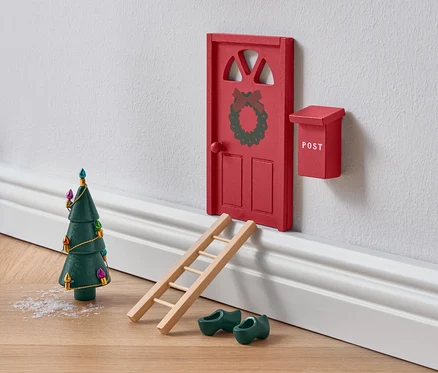 Porte rouge miniature ornée d'une couronne avec échelle, sabots en bois verts et sapin de Noël.