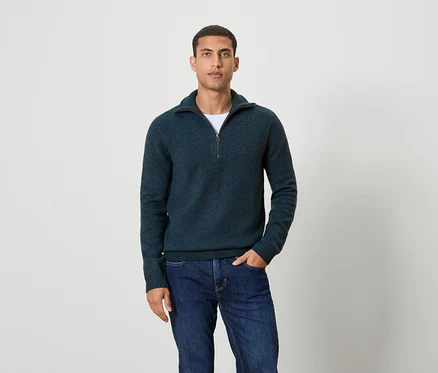 Un homme pose devant un fond portant un pull bleu foncé à fermeture éclair et un jean.