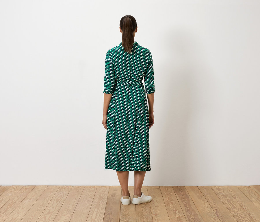 Une femme se tient dos à la caméra et porte une robe verte à motifs blancs et des baskets blanches.