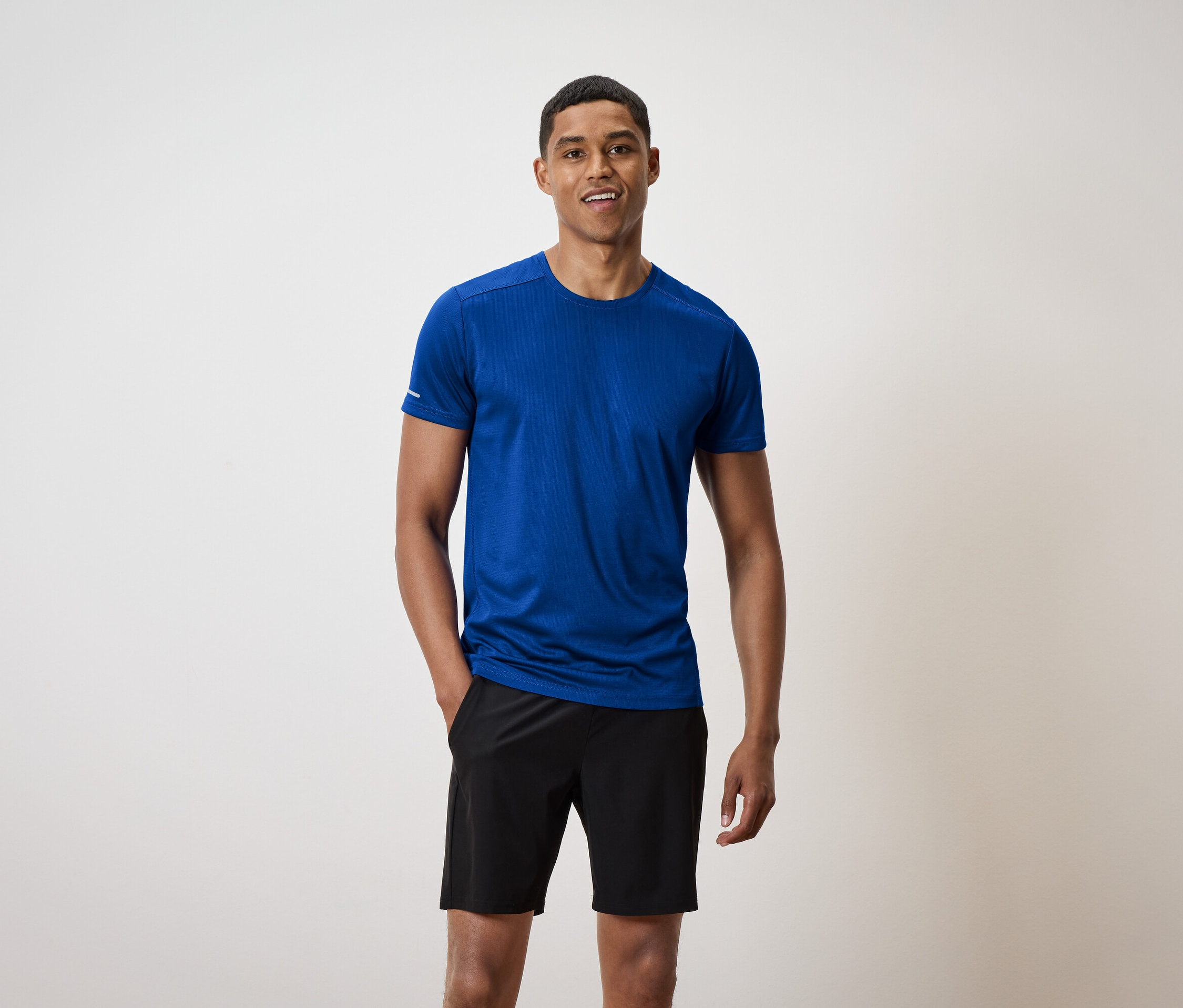 Un homme aux cheveux courts se tient debout et sourit, portant un t-shirt bleu et un short noir.