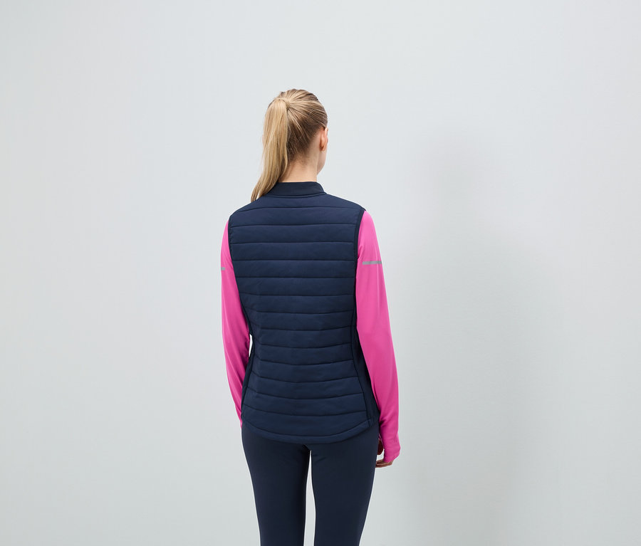 Vue arrière : femme blonde avec des cheveux attachés en queue de cheval, portant un gilet matelassé bleu marine et un T-shirt de sport rose.