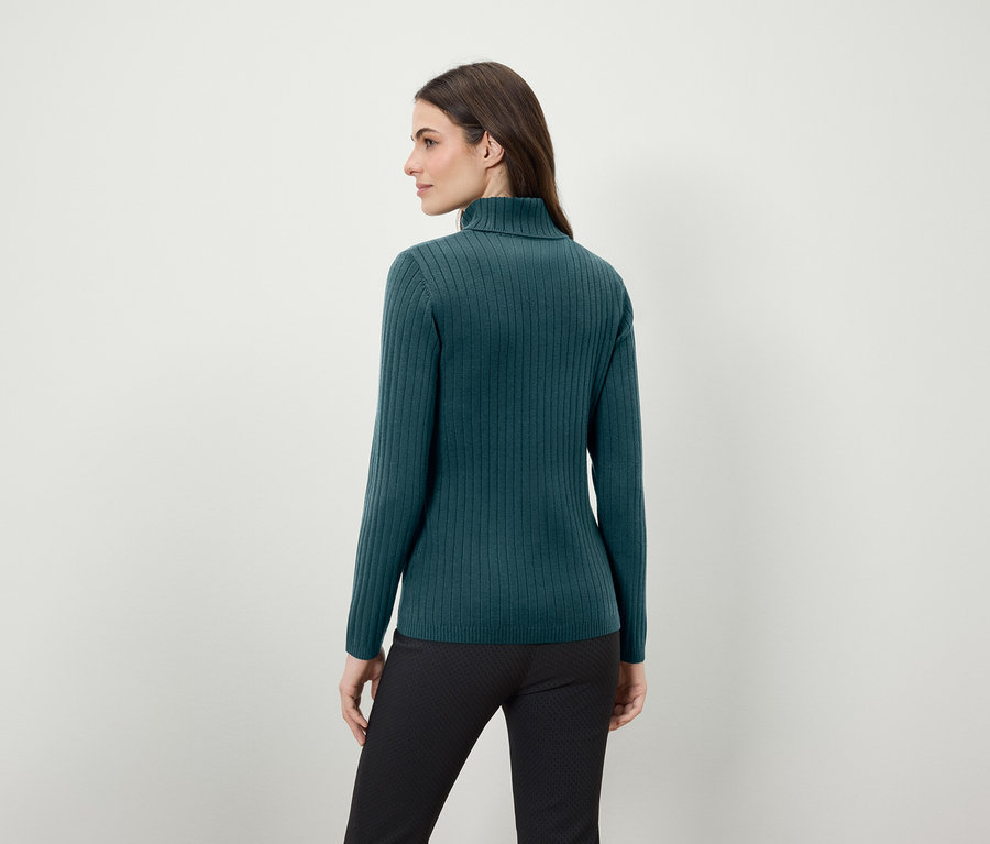 Une femme, dos à la caméra, porte un pull à col roulé vert et un pantalon noir.