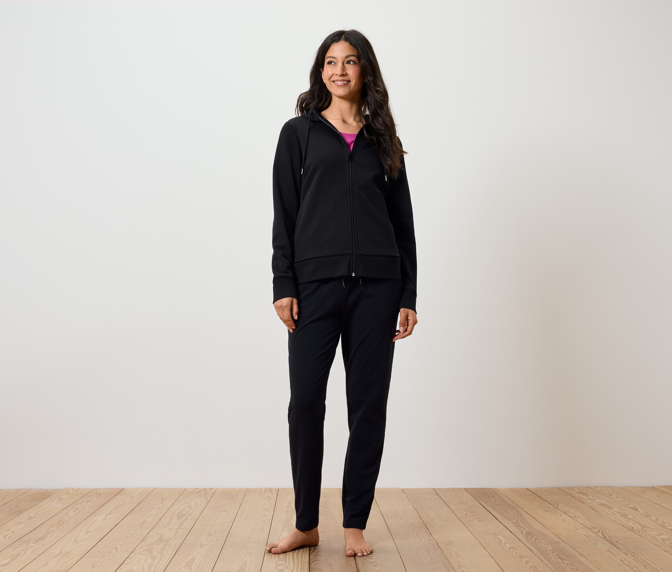 Une femme souriante se tient pieds nus sur un plancher en bois. Elle porte un pantalon noir et un sweat à capuche zippé noir.
