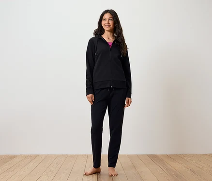 Une femme souriante se tient pieds nus sur un plancher en bois. Elle porte un pantalon noir et un sweat à capuche zippé noir.