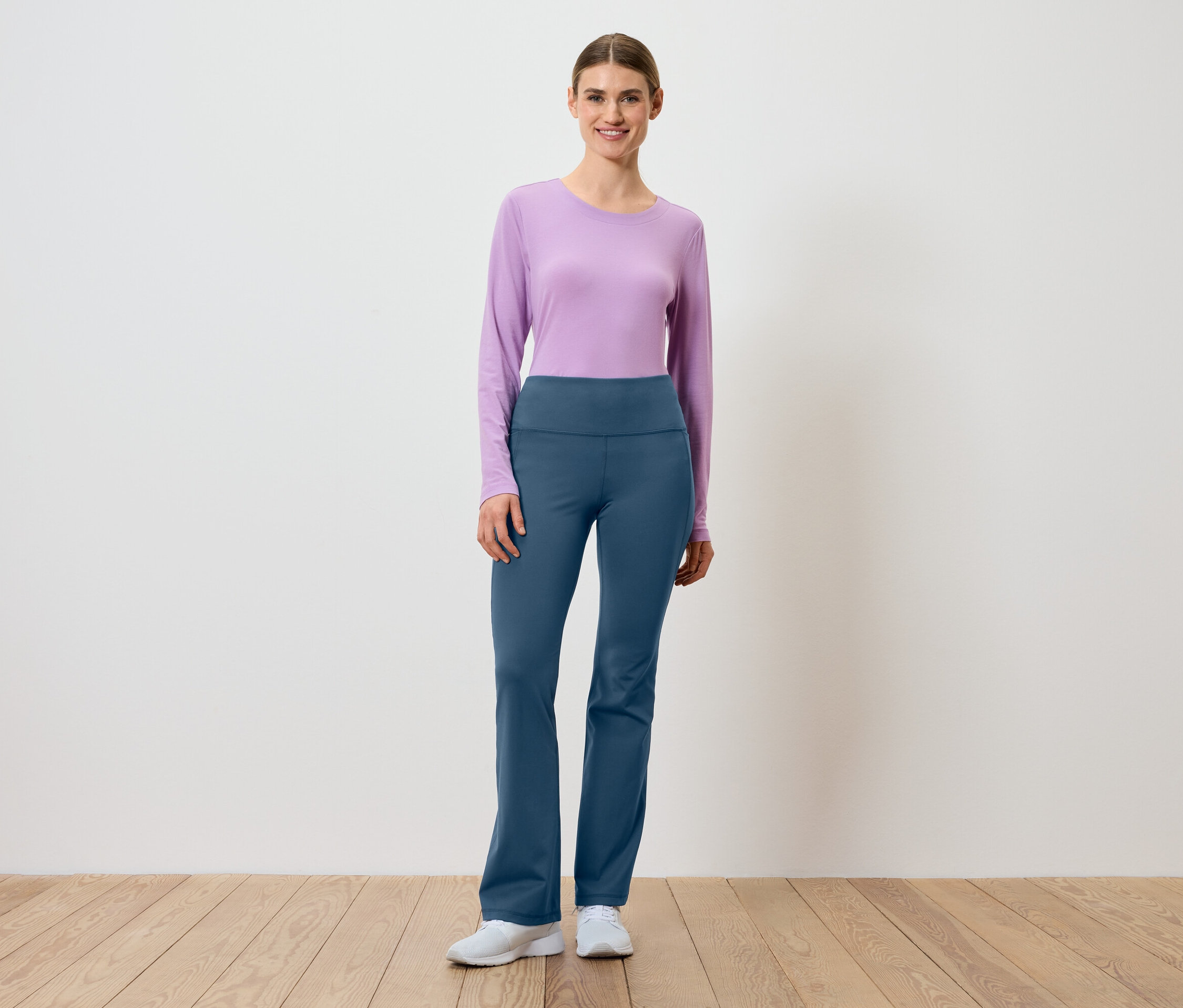 Une femme portant un t-shirt à manches longues lilas et un pantalon bleu se tient sur un plancher en bois devant un mur blanc.
