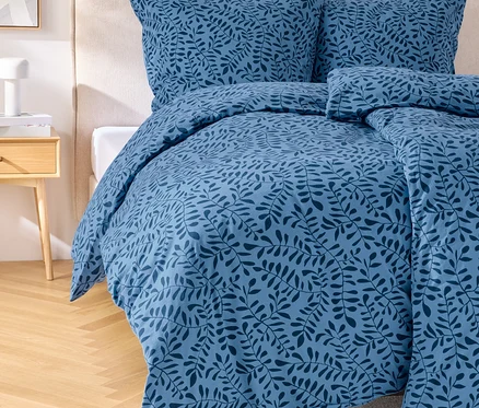 Un ensemble de literie bleu avec un motif de feuilles sur un lit avec une tête de lit. Une table de chevet avec une lampe est placée à côté du lit.