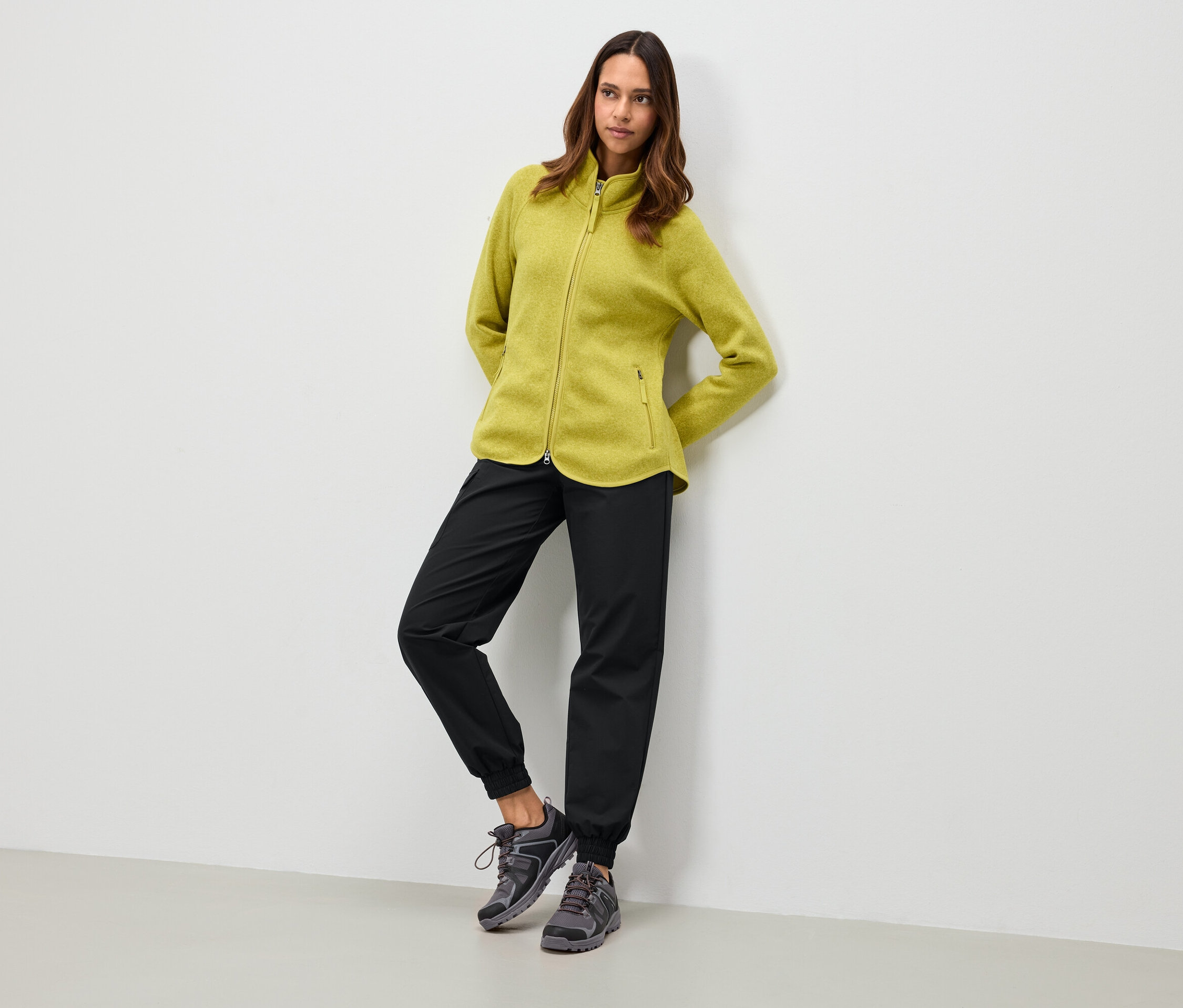 Femme portant une veste en polaire vert olive, un pantalon noir et des chaussures grises, appuyée contre un mur blanc.
