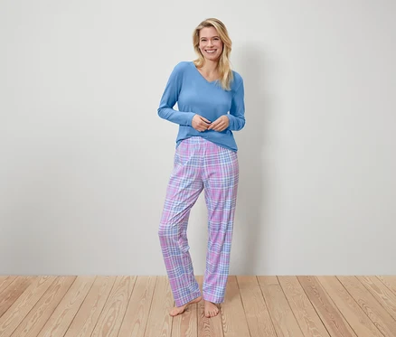 Une femme souriante se tient debout et porte un haut bleu à manches longues et un pantalon de pyjama à carreaux.
