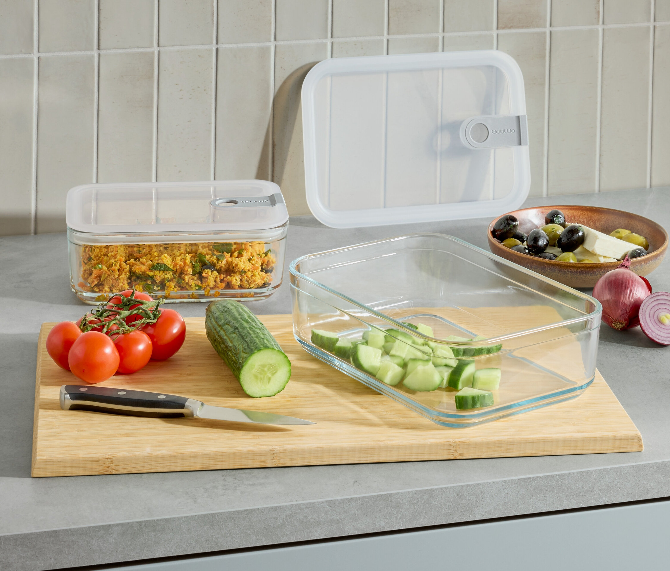 Sur un comptoir de cuisine, il y a deux récipients en verre, l'un avec de la nourriture et l'autre avec des concombres coupés. Il y a aussi des tomates, un concombre, un couteau et un bol d'olives et de fromage.