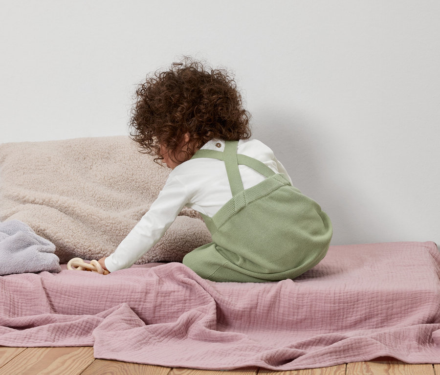 Un enfant aux cheveux bouclés agenouillé sur une couverture rose porte une salopette verte et une chemise à manches longues.
