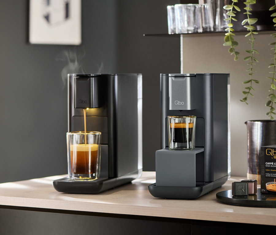 Deux machines à café sur un comptoir. La Qbo ICONIC Dark Chrome de gauche prépare du café dans un verre, tandis que la Qbo ESSENTIAL Dark Stone de droite contient déjà du café dans un verre. Des capsules Qbo by Tchibo Caffè Crema sont à côté.