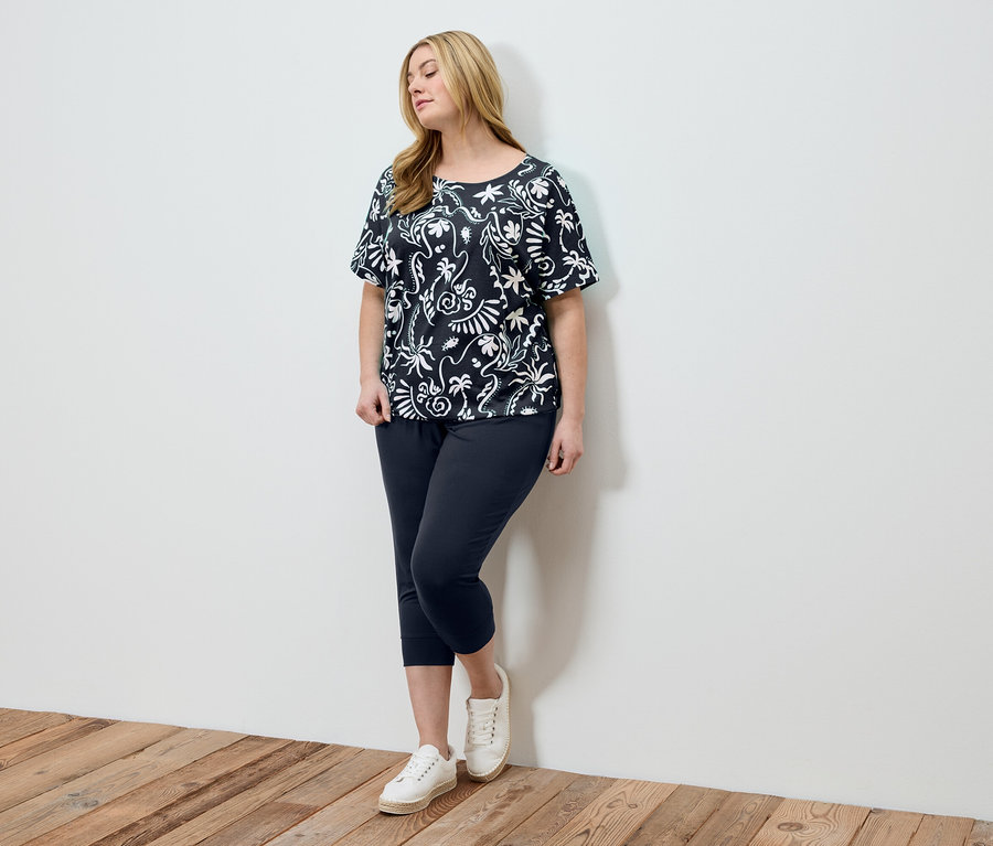 Une femme portant un haut à motifs noir et blanc, un pantalon foncé et des chaussures blanches se tient devant un mur blanc.