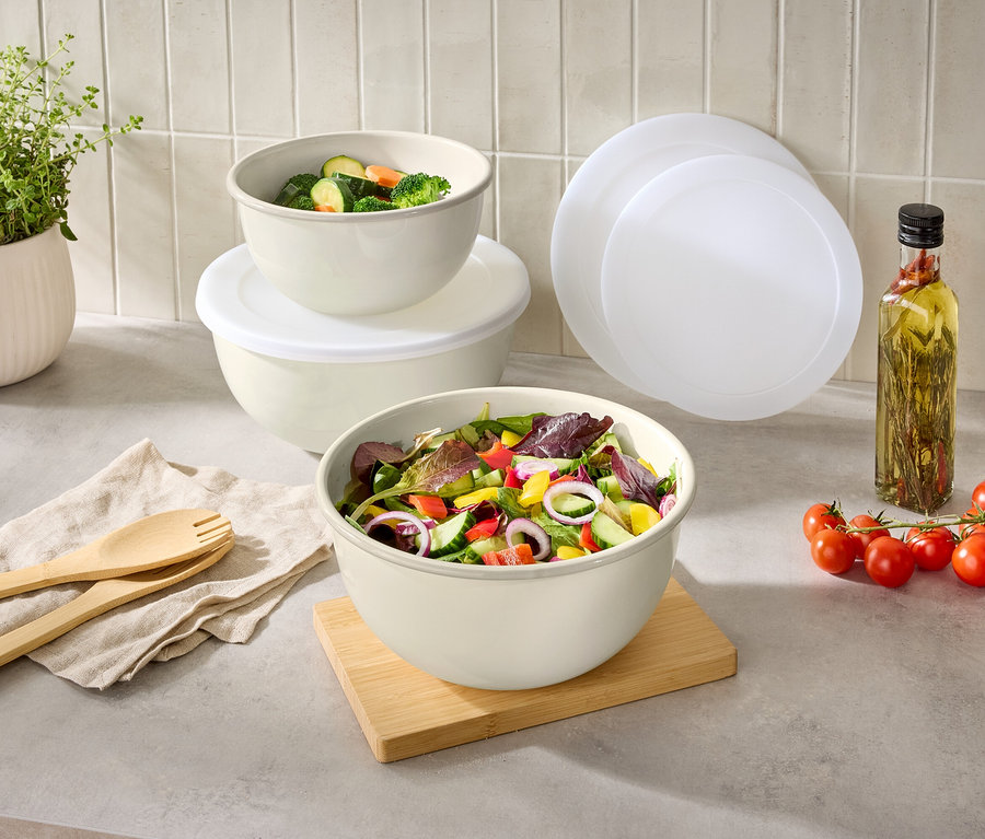 Sur un plan de travail de cuisine, des saladiers avec de la salade et des légumes, à côté d'un pot d'herbes aromatiques, des couverts à salade, des tomates, de l'huile et des couvercles.