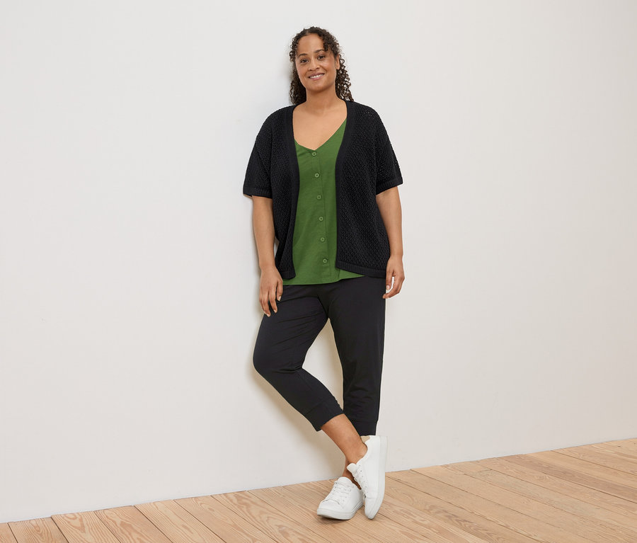 Une femme portant un cardigan en maille noire sur une chemise vert olive, un pantalon noir et des baskets blanches est appuyée contre un mur blanc.