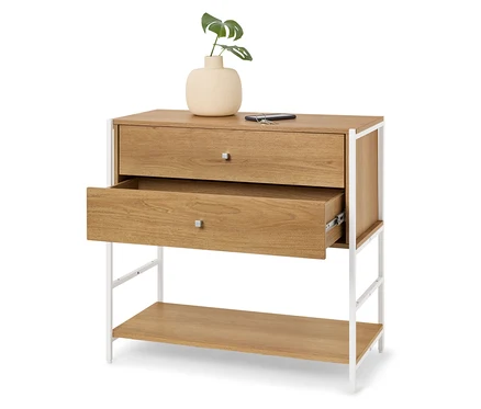 Commode en bois clair avec deux tiroirs, dont un est ouvert, et un cadre en métal blanc. Un vase avec une plante verte est posé sur le dessus.