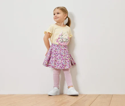Une petite fille blonde porte un t-shirt à rayures jaunes avec un motif floral, une jupe à fleurs, un legging rose et des baskets blanches.