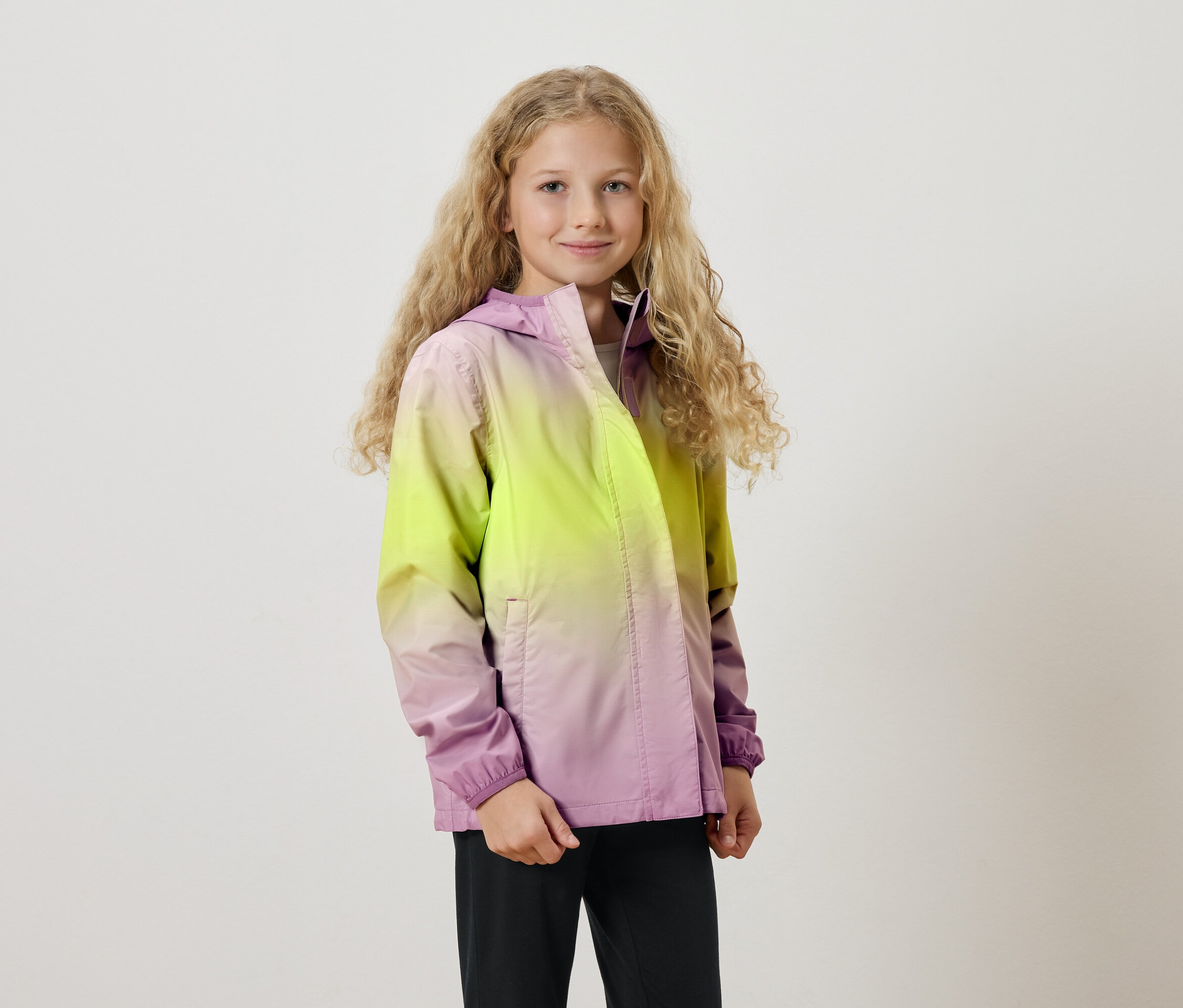 Une fille aux longs cheveux blonds bouclés porte une veste à capuche avec un dégradé de couleurs allant du jaune au violet.