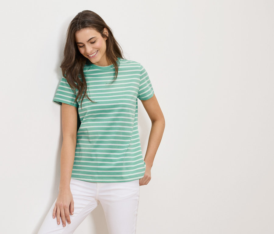 Une femme aux cheveux bruns porte un t-shirt rayé vert et blanc et un pantalon blanc.