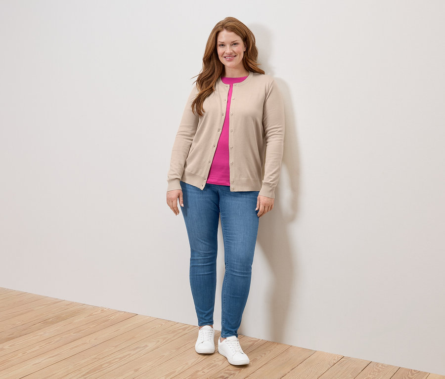 Une femme aux longs cheveux roux se tient devant un mur, portant un cardigan beige, un t-shirt rose, un jean et des baskets blanches.
