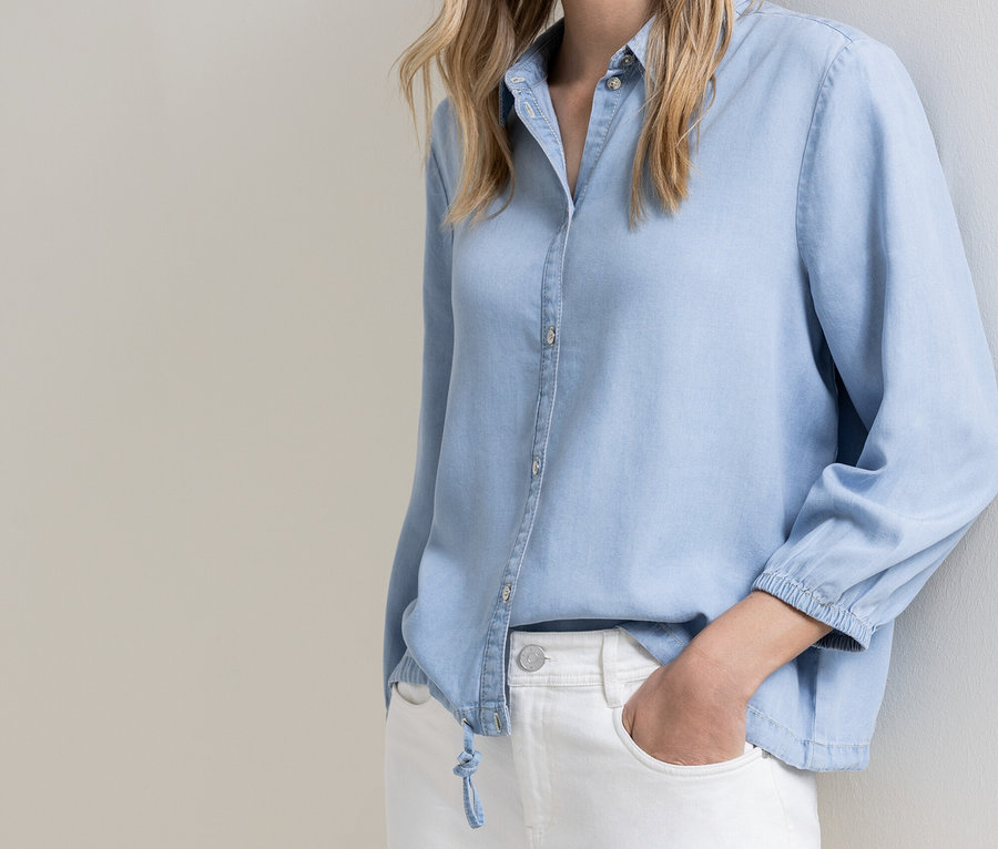 Une femme portant une chemise boutonnée bleu clair et un jean blanc, une main dans la poche, appuyée contre un mur.
